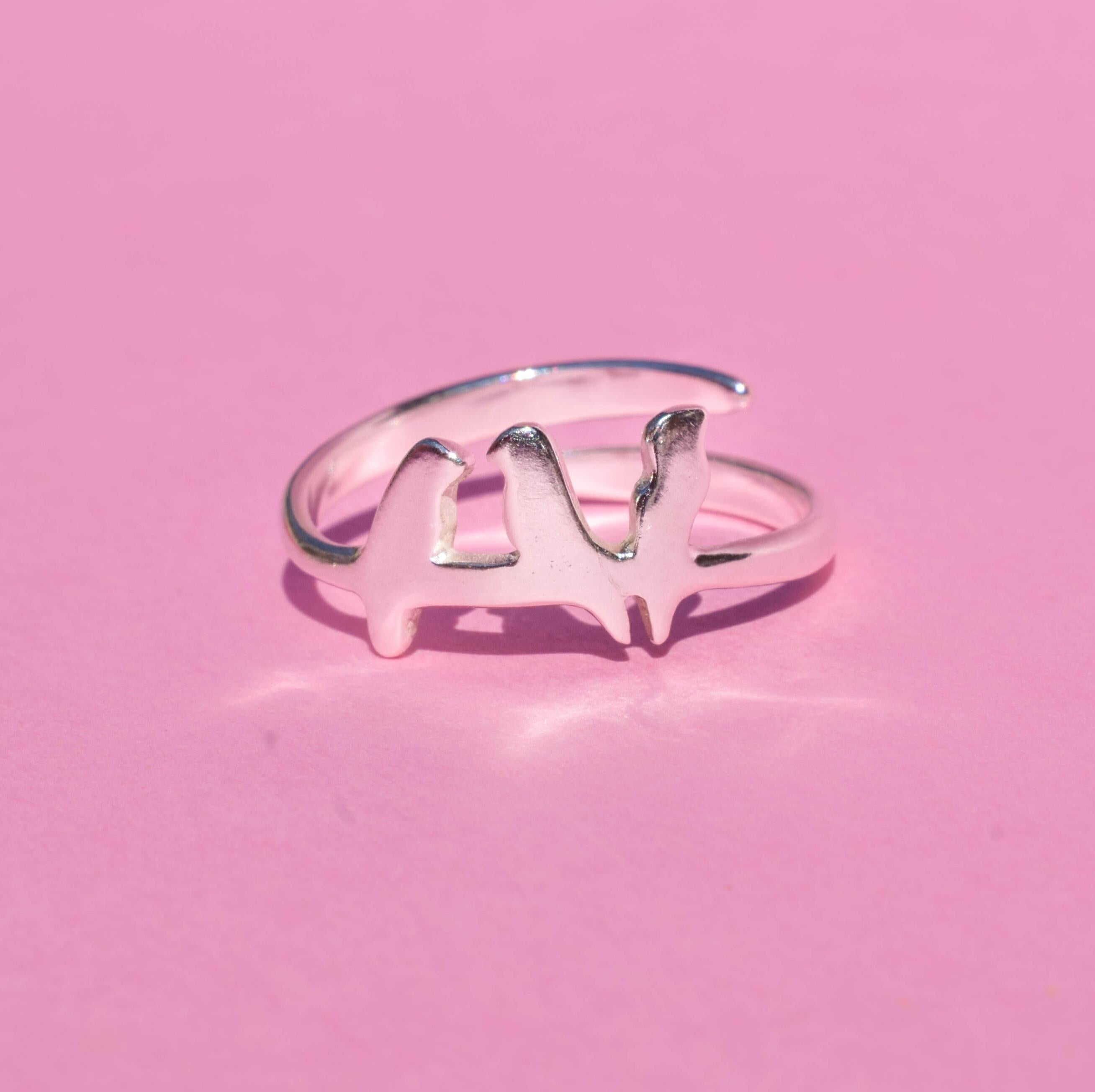 A silver ring with three bird designs perched on a wire, displayed against a pink background.