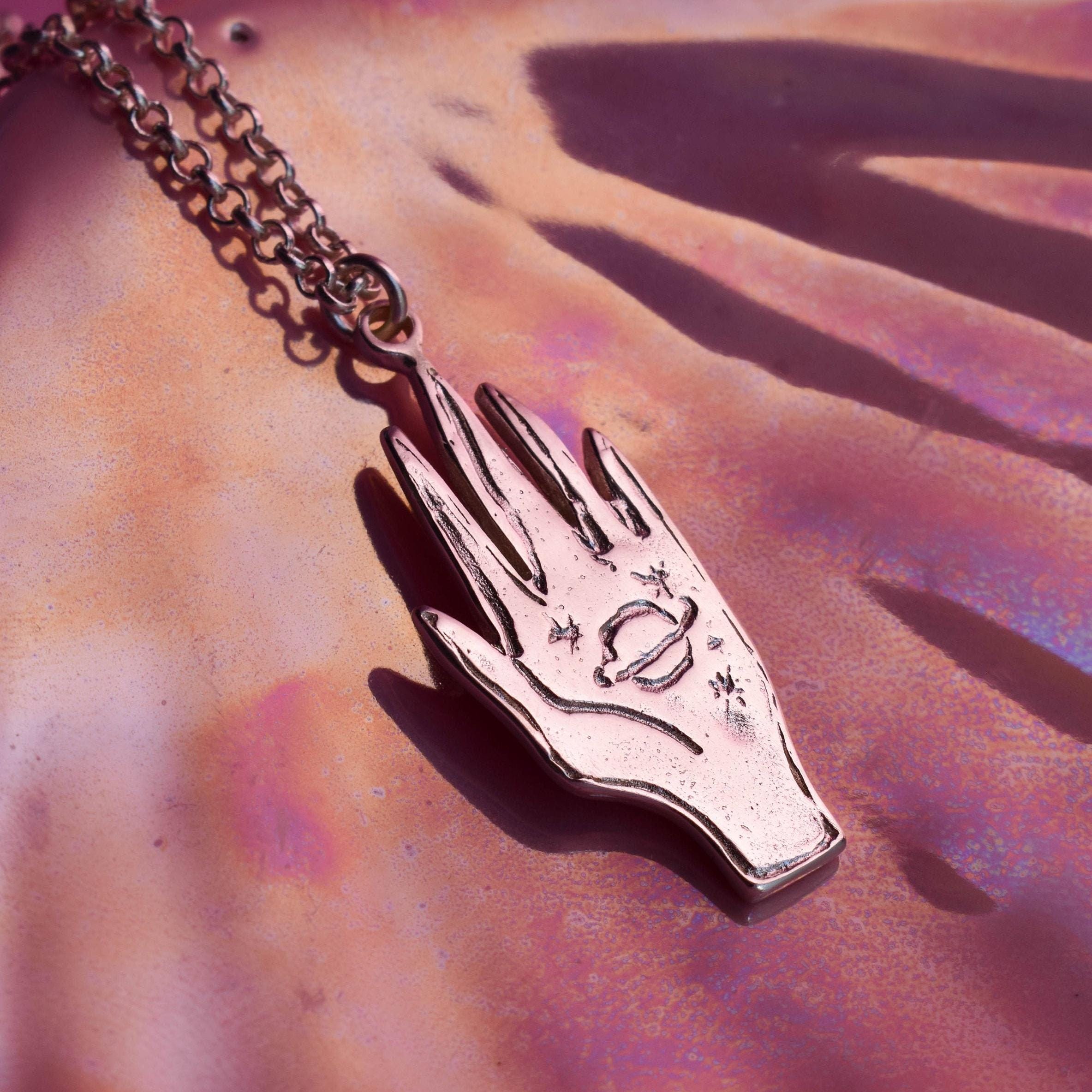 A silver pendant in the shape of a hand with etched celestial stars, attached to a chain, displayed against a patterned background.