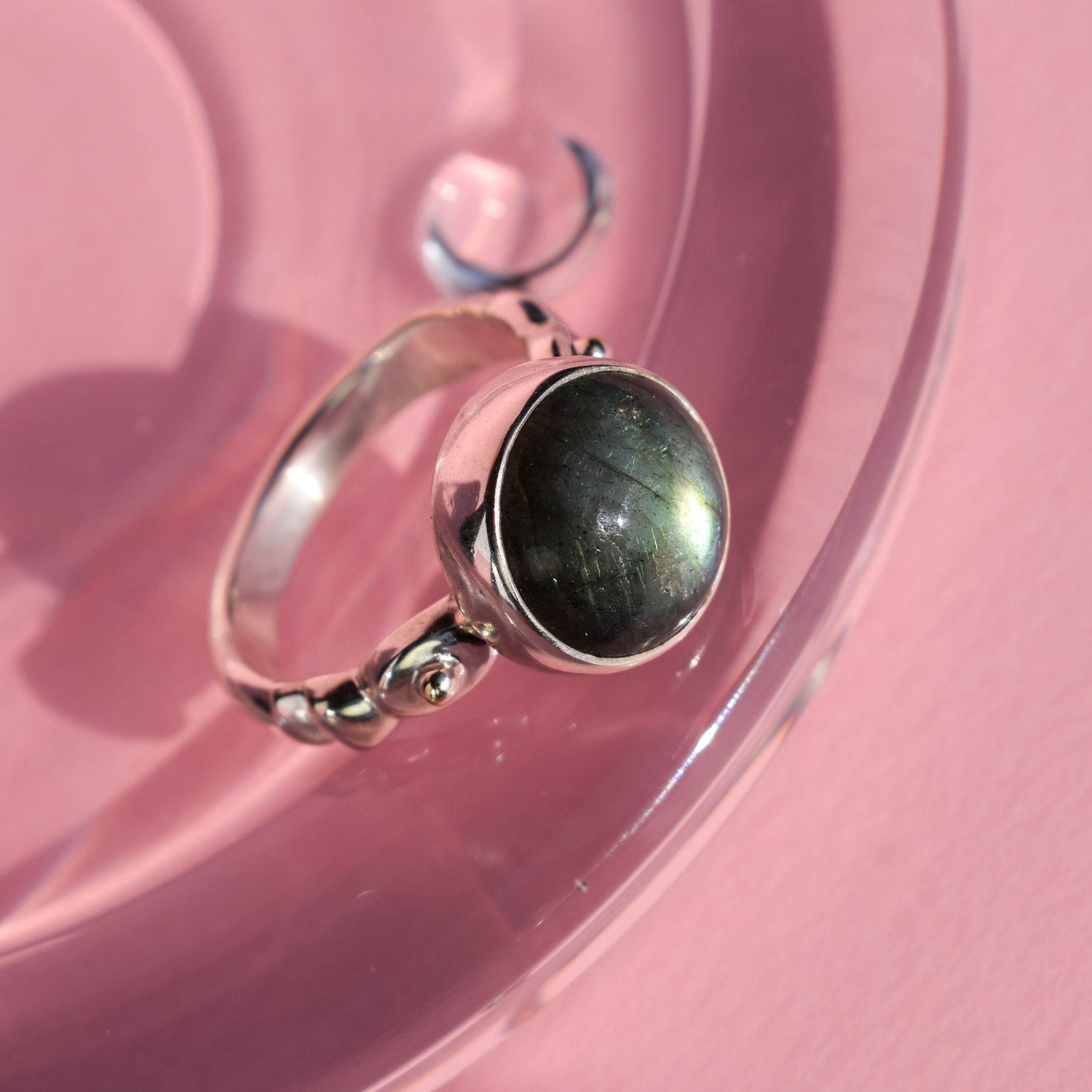 A silver ring with a large oval dark gray stone, resting on a glossy pink surface.