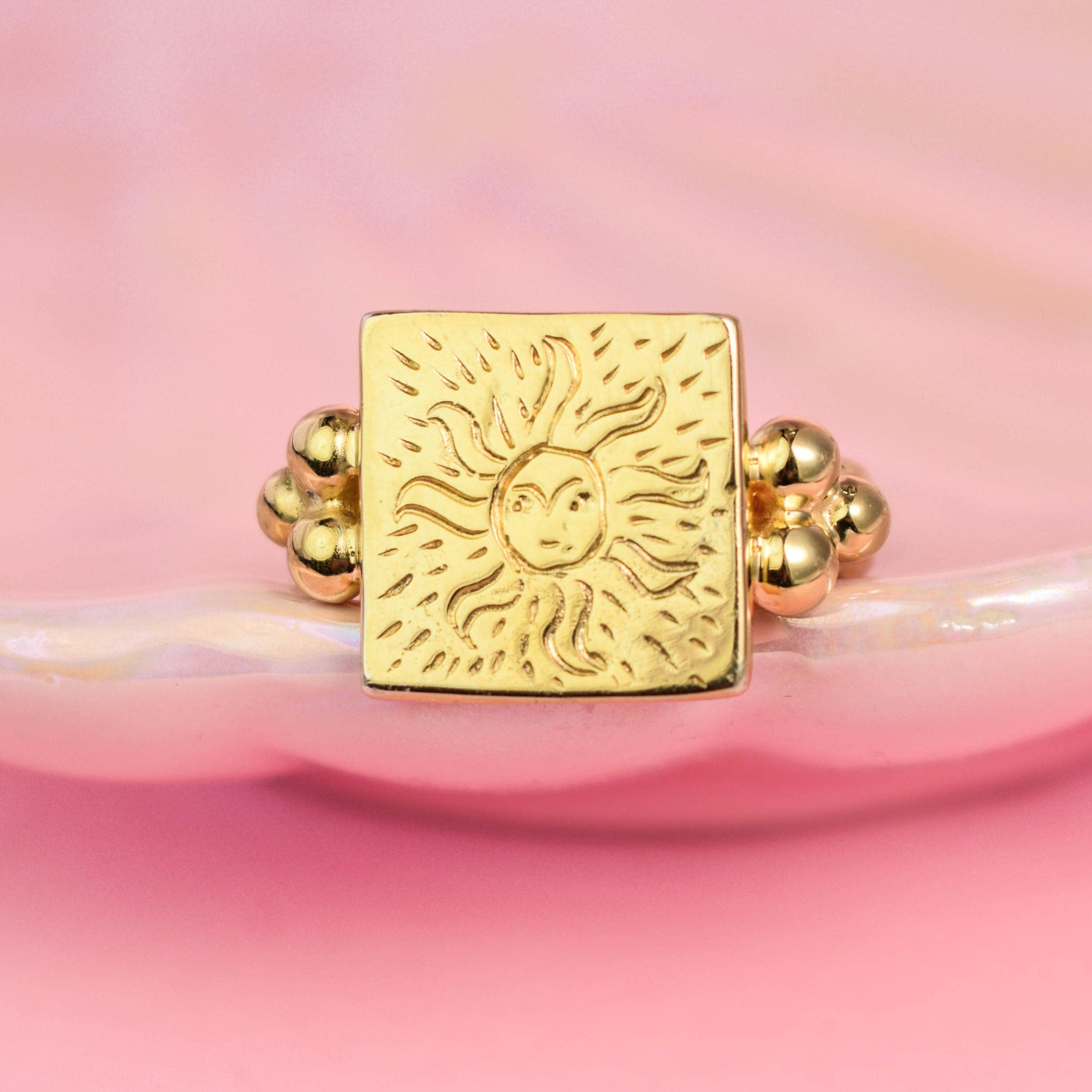 A square gold-colored ring with a sun engraving on a pink background